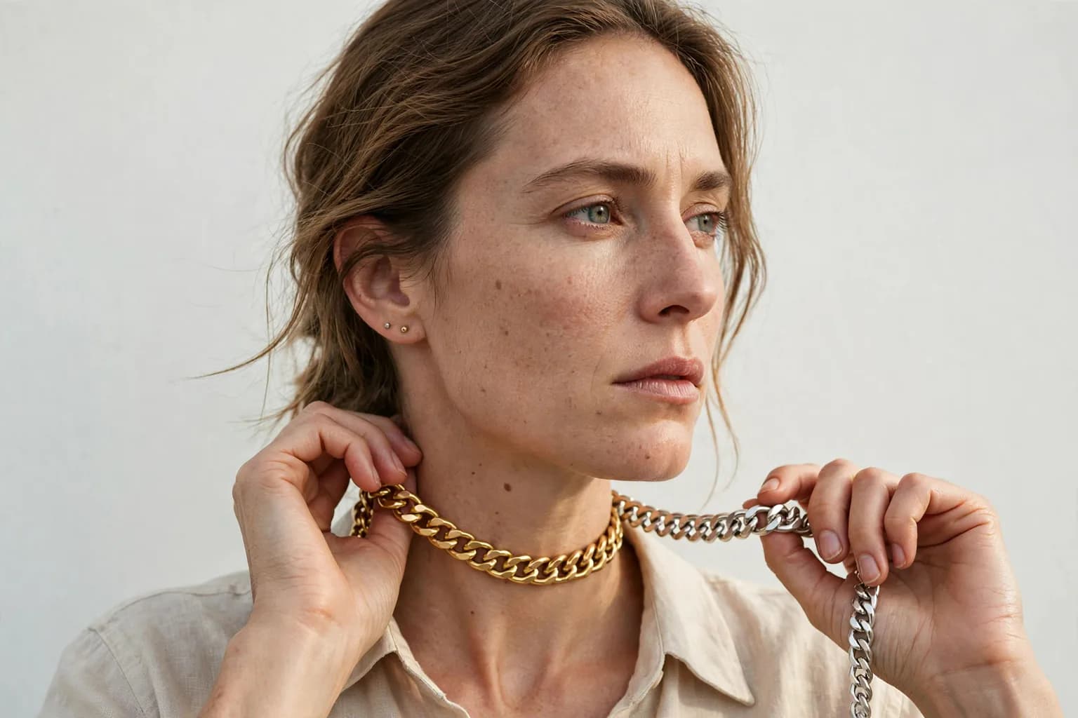 A woman holding a gold chain necklace at her neck and a silver chain in her other hand, comparing both metals against her skin in daylight
